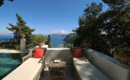 Luxurious outdoor seating area with two sun loungers and a small table overlooking a stunning coastal view, surrounded by lush greenery and a swimming pool.