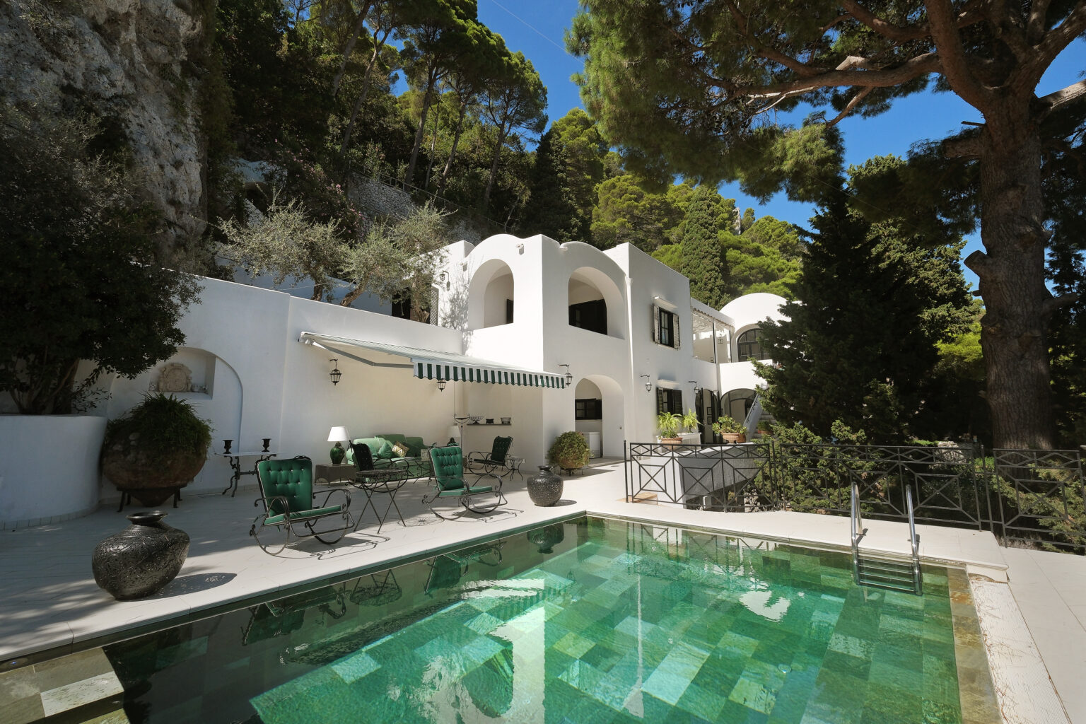 Luxurious white villa with a swimming pool, surrounded by lush greenery and tall trees, showcasing a modern architectural design and outdoor seating area for relaxation. | Only on SopranoVillas