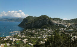 Scenic view of Capri, Italy, showcasing lush greenery, coastal cliffs, and a vibrant blue sea under a clear sky, with boats anchored in the harbor and charming white buildings nestled on the hillside.