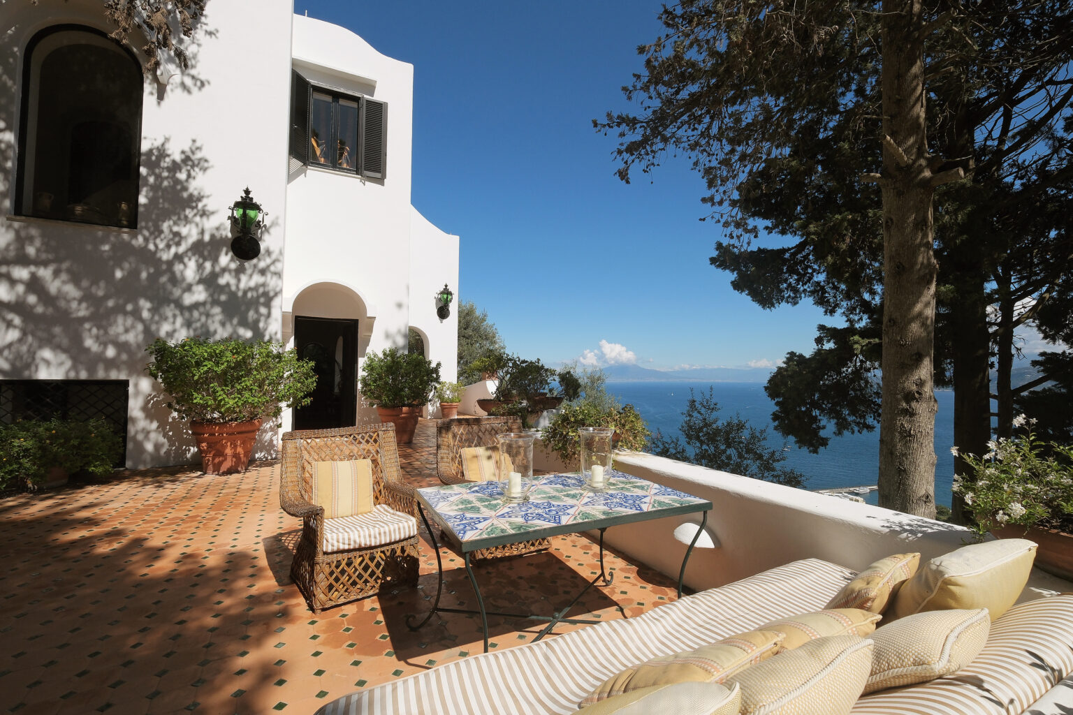 Mediterranean villa terrace with ocean view, featuring a dining table, comfortable seating, and lush greenery, under a clear blue sky. Ideal for outdoor relaxation and entertaining. | Only on SopranoVillas