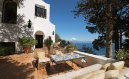Mediterranean villa terrace with ocean view, featuring a dining table, comfortable seating, and lush greenery, under a clear blue sky. Ideal for outdoor relaxation and entertaining.
