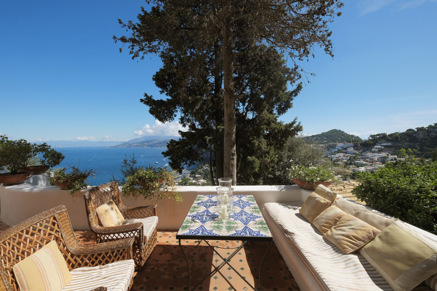 Cozy outdoor seating area with a colorful tiled table, surrounded by lush greenery and offering a stunning view of the sea and distant hills on a sunny day. | Only on SopranoVillas