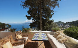 Cozy outdoor seating area with a colorful tiled table, surrounded by lush greenery and offering a stunning view of the sea and distant hills on a sunny day.