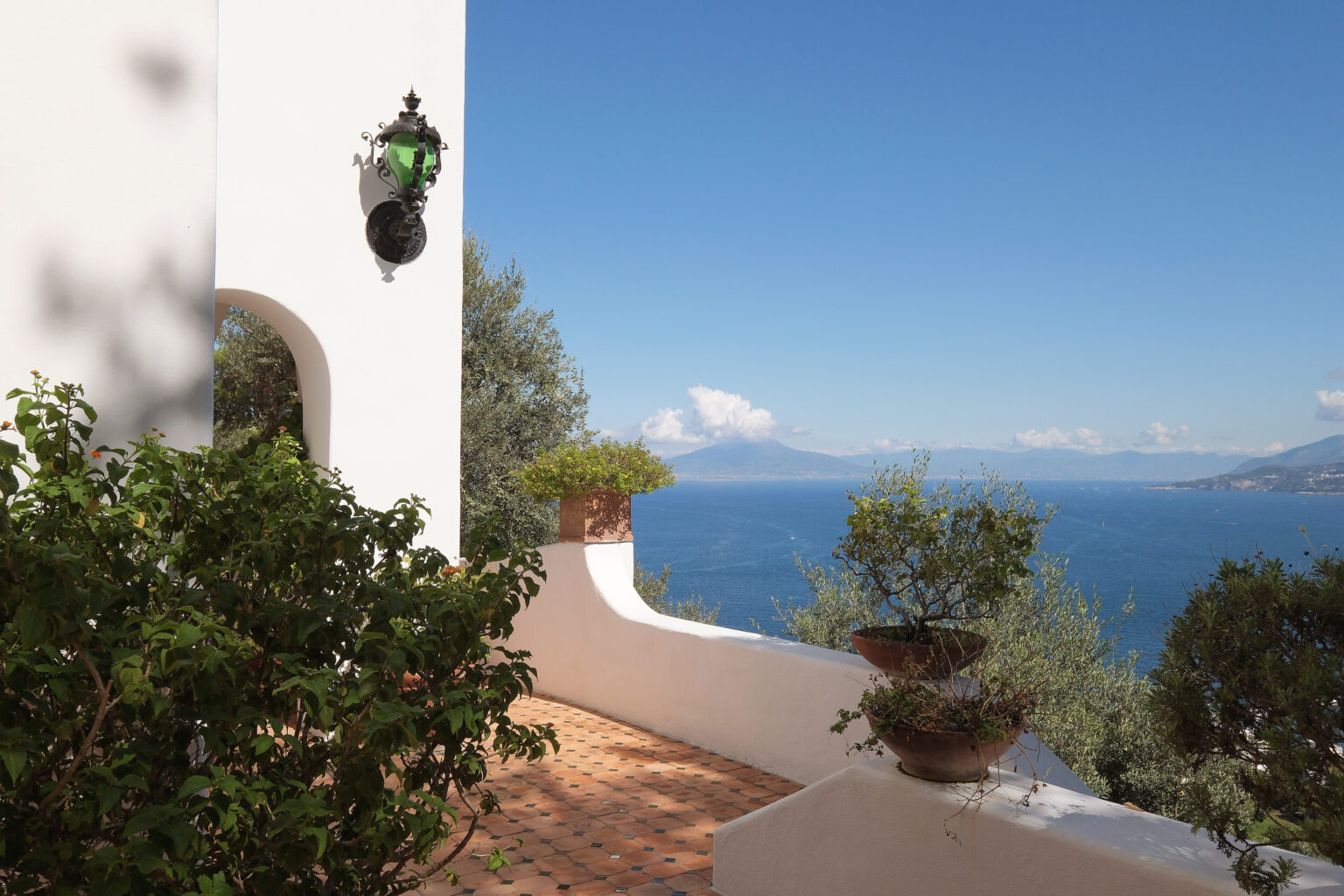Scenic view from a terrace overlooking a tranquil sea, surrounded by lush greenery and a charming white wall, with a decorative lantern adding to the Mediterranean ambiance. | Only on SopranoVillas
