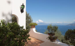 Scenic view from a terrace overlooking a tranquil sea, surrounded by lush greenery and a charming white wall, with a decorative lantern adding to the Mediterranean ambiance.