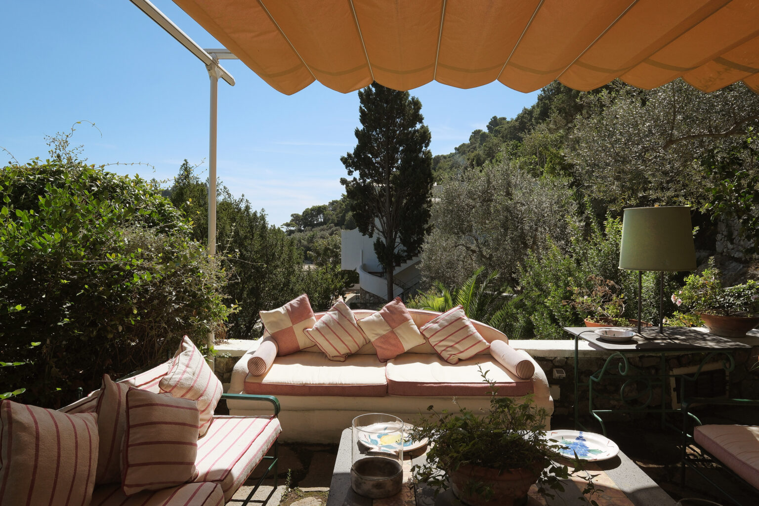 Cozy outdoor seating area with plush cushions under a yellow awning, surrounded by lush greenery and a distant view of a hillside. Ideal for relaxation and enjoying nature. | Only on SopranoVillas