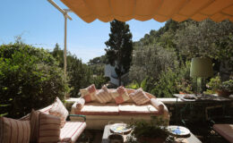Cozy outdoor seating area with plush cushions under a yellow awning, surrounded by lush greenery and a distant view of a hillside. Ideal for relaxation and enjoying nature.