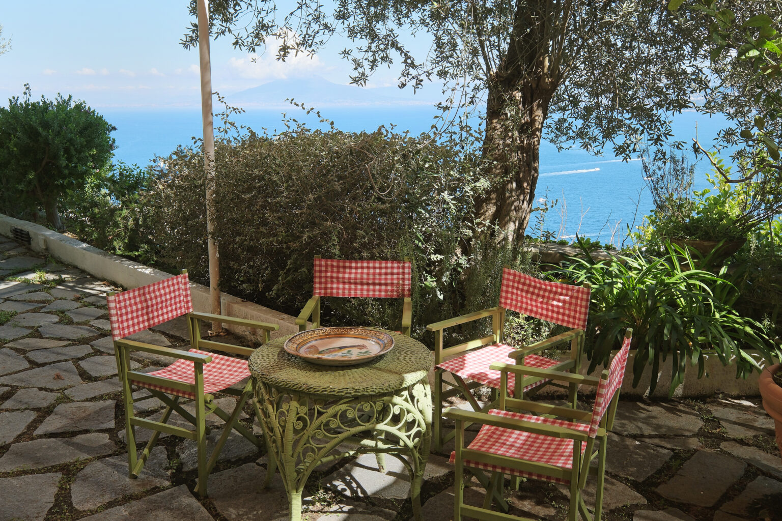 Outdoor seating area with four vintage-style chairs and a round table, surrounded by lush greenery and a scenic view of the ocean. Perfect for relaxation or dining al fresco in a tranquil setting. | Only on SopranoVillas
