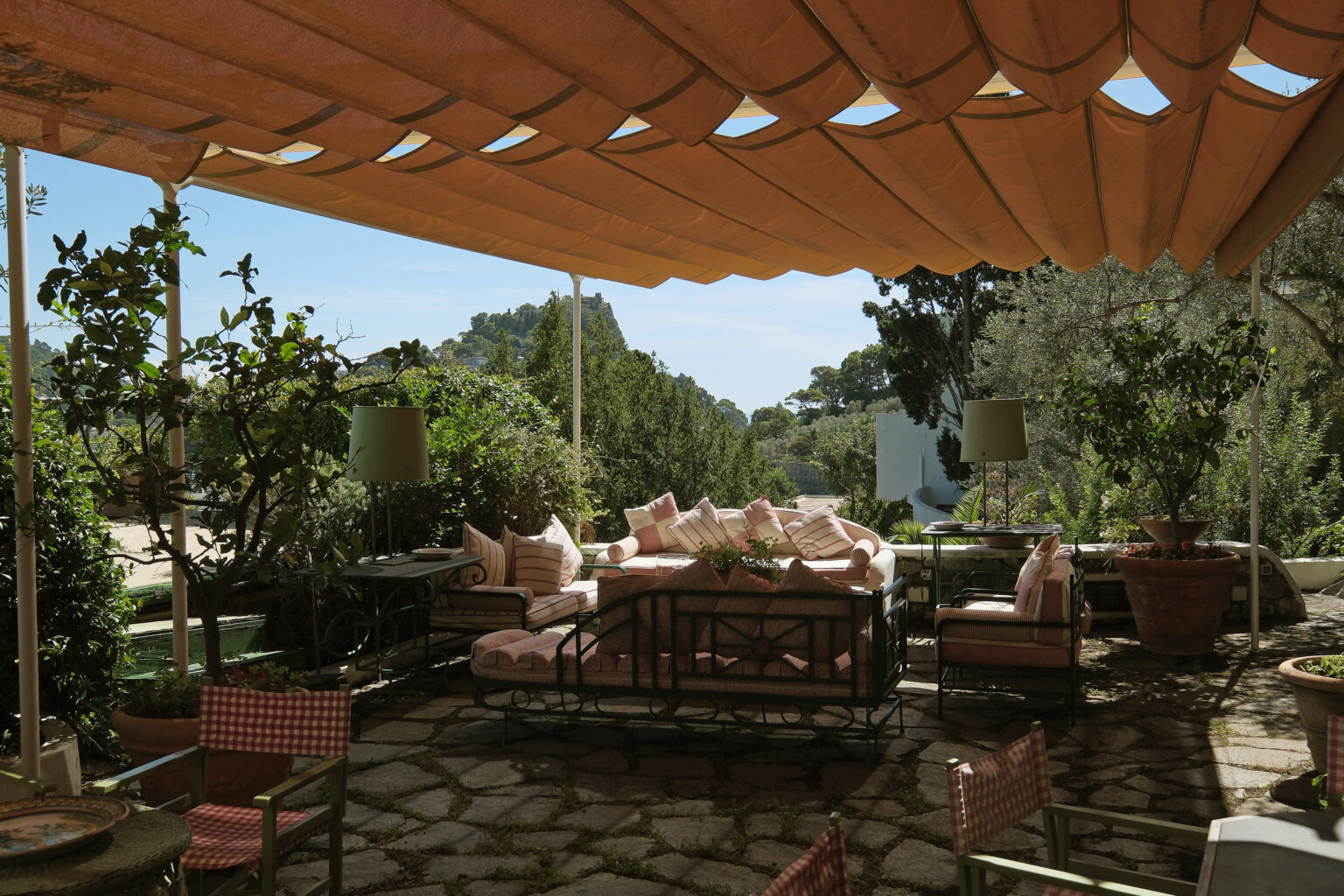 Cozy outdoor living space featuring a shaded area with a large sofa adorned with pink cushions, surrounded by greenery and decorative plants, ideal for relaxation and entertaining. | Only on SopranoVillas