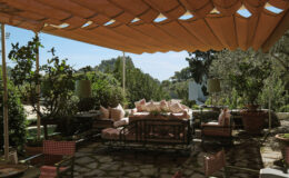 Cozy outdoor living space featuring a shaded area with a large sofa adorned with pink cushions, surrounded by greenery and decorative plants, ideal for relaxation and entertaining.
