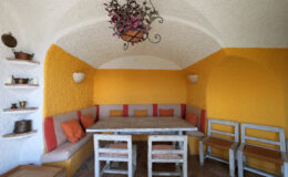 Cozy interior of a dining area featuring a rustic table and comfortable seating with orange and neutral tones, accented by decorative wall shelves and a stylish chandelier. Ideal for gatherings and relaxation.
