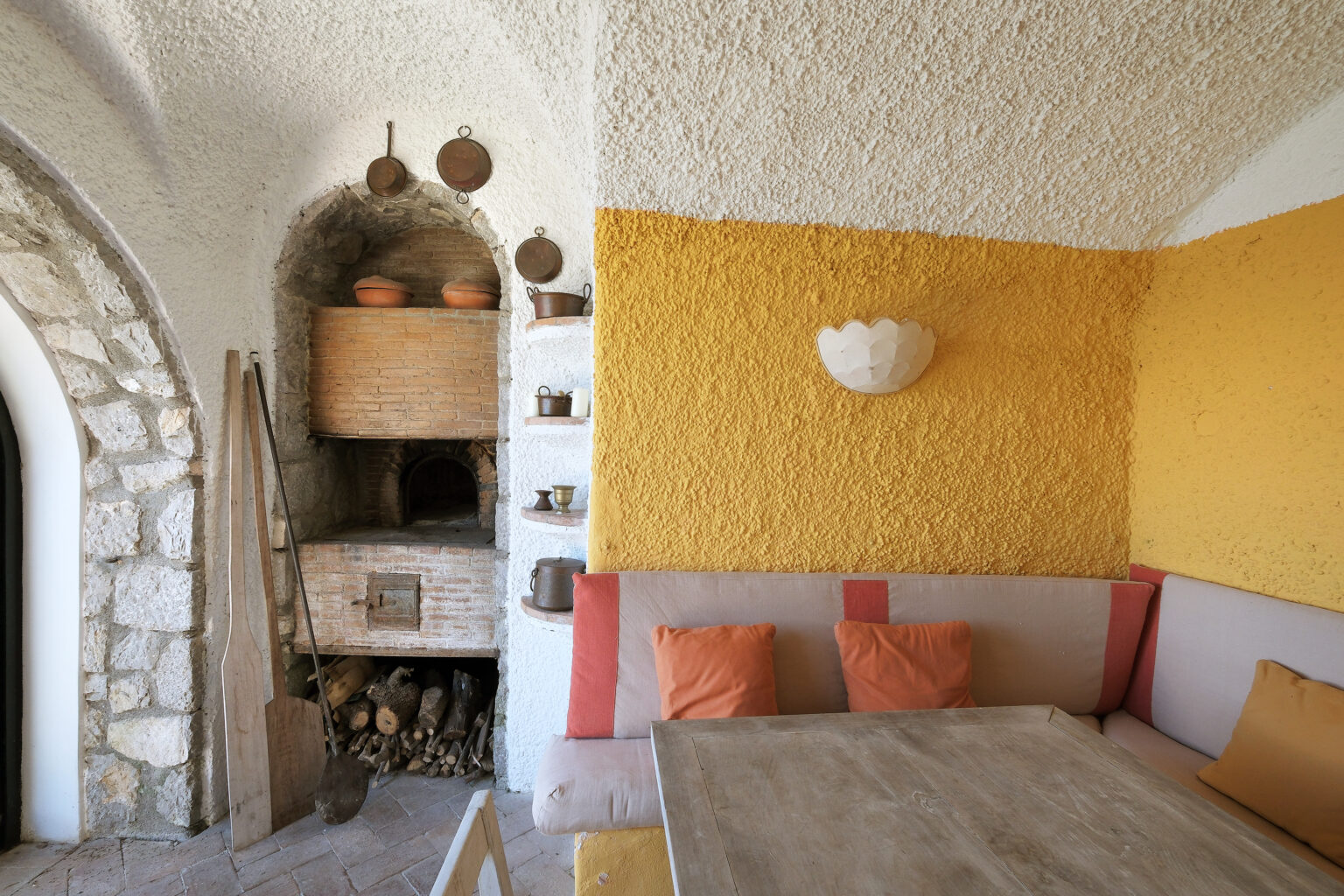 Cozy interior of a rustic room featuring a stone wall, a wooden dining table, and a comfortable seating area adorned with orange cushions. The wall is painted in a warm yellow hue, and a traditional oven is visible in the background, adding to the inviting atmosphere. | Only on SopranoVillas