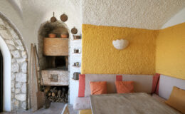 Cozy interior of a rustic room featuring a stone wall, a wooden dining table, and a comfortable seating area adorned with orange cushions. The wall is painted in a warm yellow hue, and a traditional oven is visible in the background, adding to the inviting atmosphere.