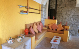 Cozy living room with a yellow wall, featuring a comfortable sofa adorned with colorful cushions, a stone wall backdrop, decorative teapots, and a small coffee table.