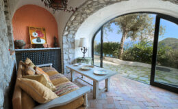 Cozy interior of a rustic living room featuring a stone archway, comfortable sofa with decorative pillows, a wooden coffee table, and large glass doors opening to a scenic outdoor view with lush greenery.