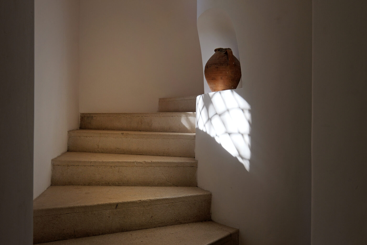 A serene staircase with natural stone steps, illuminated by soft sunlight filtering through a window, showcasing a decorative clay pot at the top. | Only on SopranoVillas