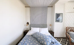Cozy bedroom featuring a double bed with blue floral bedding, soft lighting from bedside lamps, and a minimalist aesthetic. The room has a light-colored wall and a decorative wall hanging, creating a serene atmosphere. Ideal for relaxation and comfort.