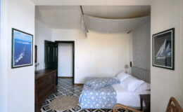 Cozy bedroom featuring a double bed with blue and white bedding, wooden furniture, and nautical-themed artwork on the walls. The room is well-lit, showcasing a blend of modern and classic design elements.