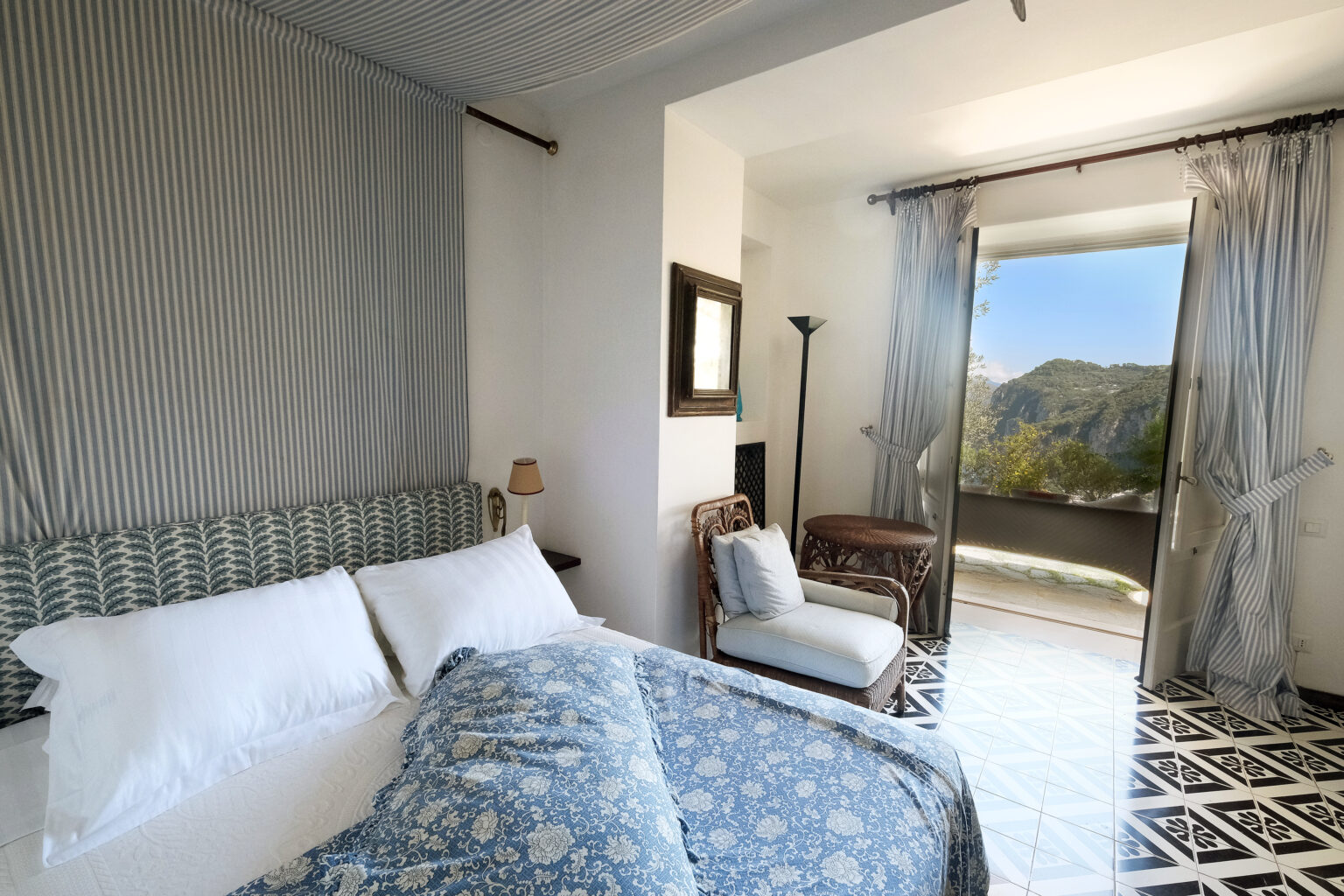 Cozy bedroom featuring a patterned blue bedspread, white linens, and a comfortable armchair, with large windows showcasing a scenic outdoor view of mountains. Bright and airy design with light-colored walls and elegant curtains. | Only on SopranoVillas
