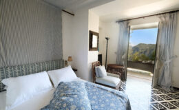 Cozy bedroom featuring a patterned blue bedspread, white linens, and a comfortable armchair, with large windows showcasing a scenic outdoor view of mountains. Bright and airy design with light-colored walls and elegant curtains.