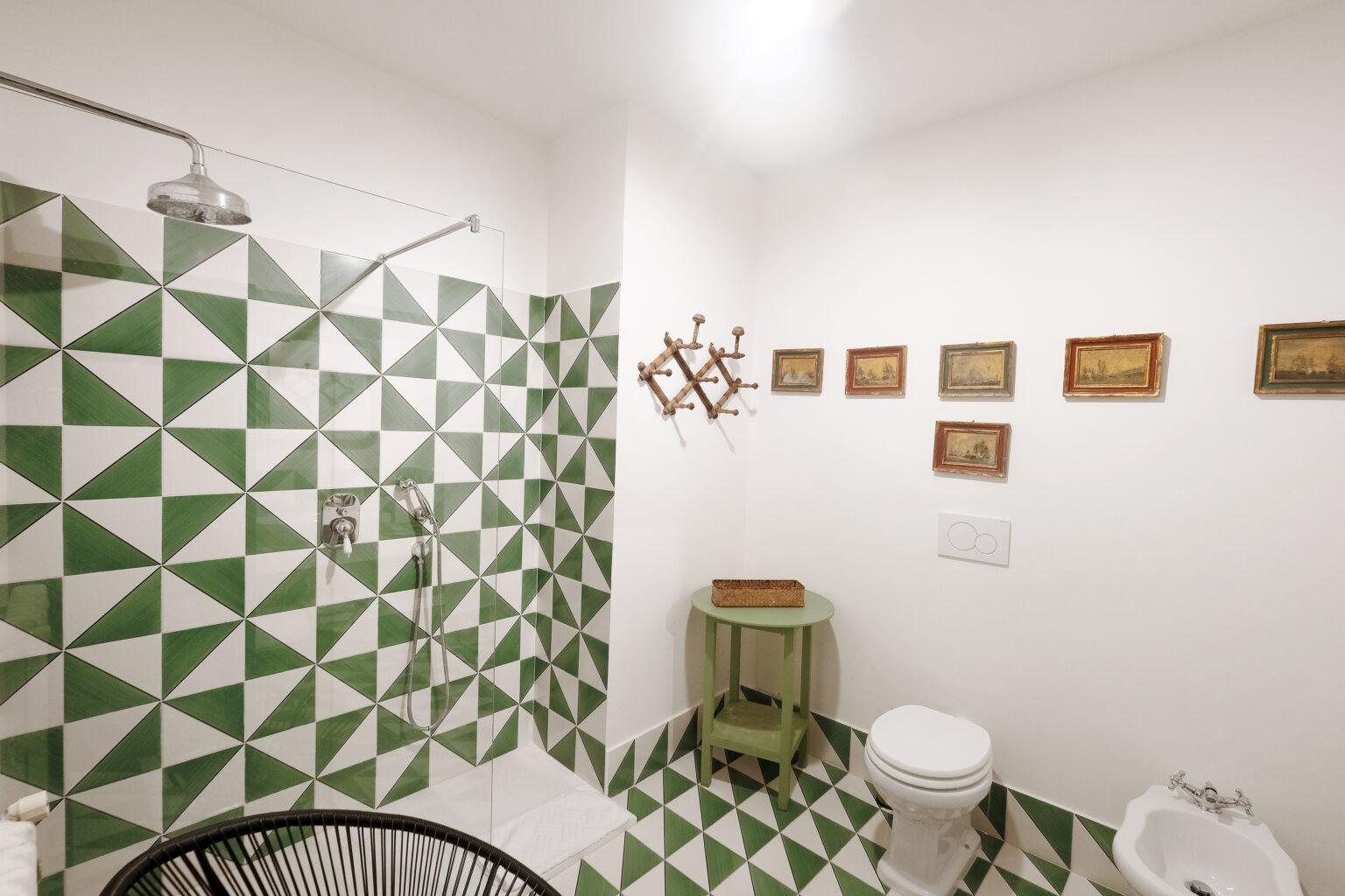 Modern bathroom featuring a unique green and white geometric tile design, a shower with a sleek fixture, a compact toilet, and decorative wall art. A small green side table adds a touch of color to the space. | Only on SopranoVillas