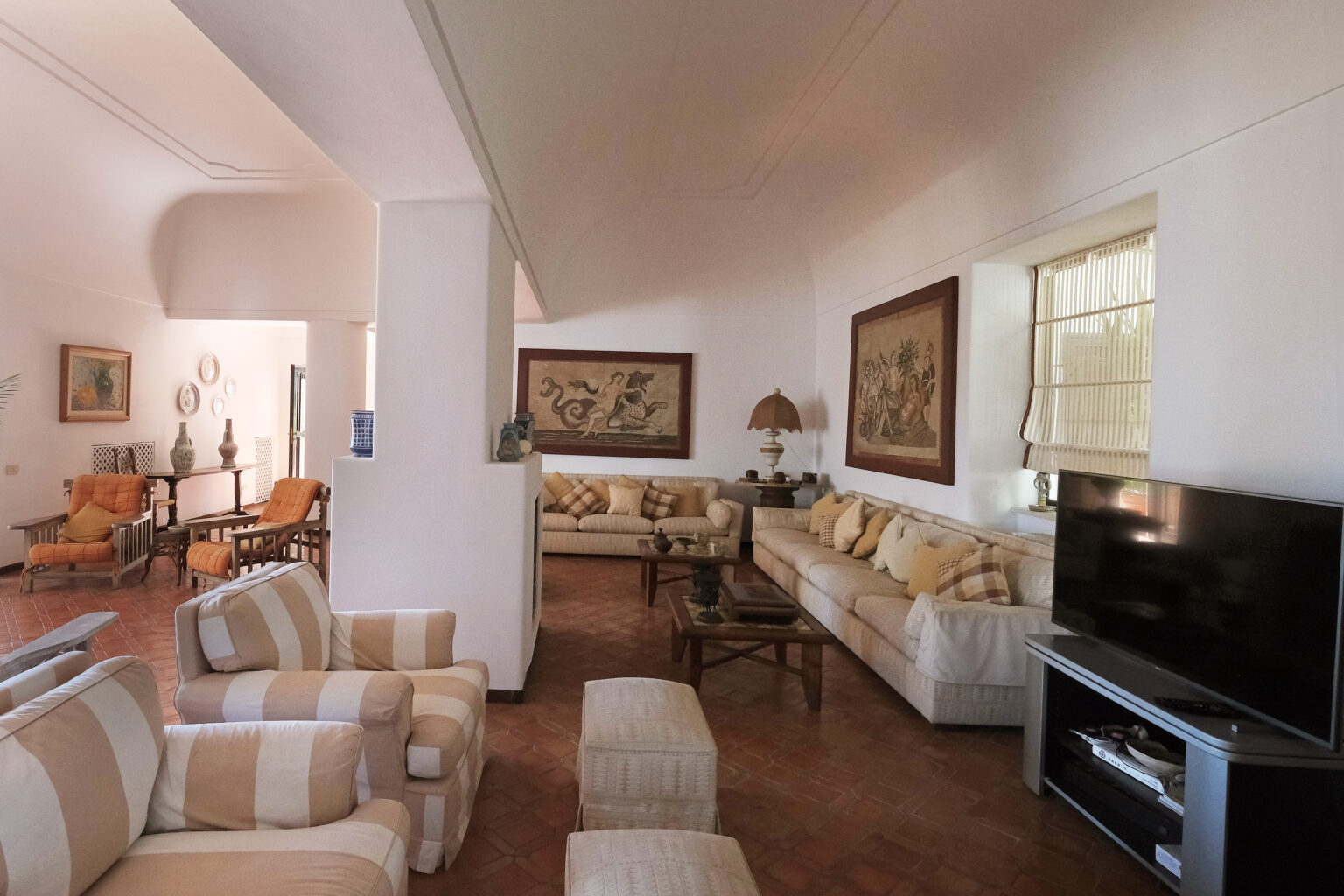 Spacious living room featuring comfortable seating, decorative paintings, and a modern television, with natural light enhancing the warm atmosphere. | Only on SopranoVillas