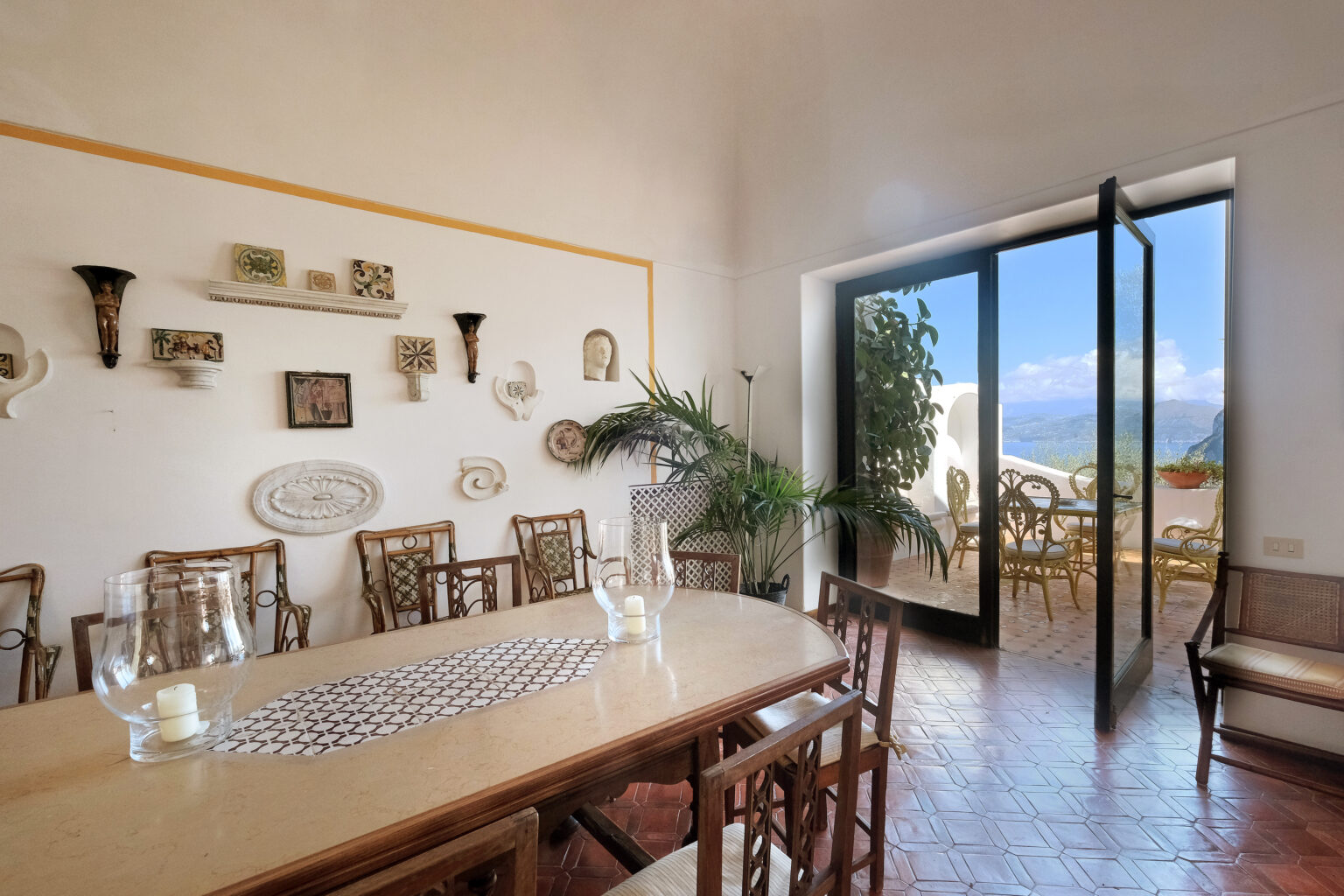 Dining room with a large wooden table, surrounded by elegant chairs, featuring decorative wall plates and a view of the outdoor terrace through open glass doors, showcasing a scenic landscape. | Only on SopranoVillas