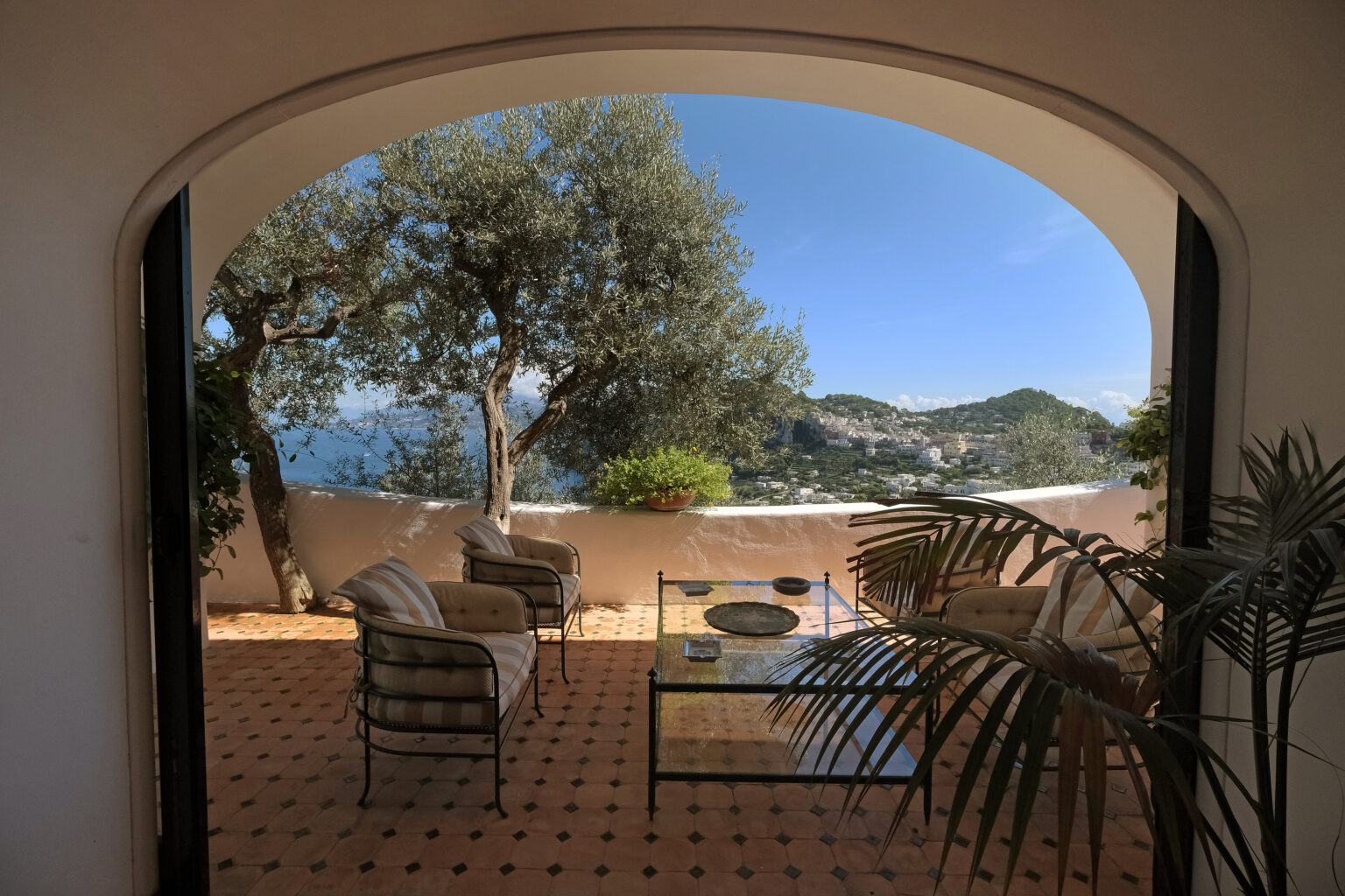 Cozy outdoor seating area with a glass table, overlooking a scenic view of hills and coastline, framed by an arched entrance and olive trees. Ideal for relaxation and enjoying nature. | Only on SopranoVillas