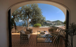 Cozy outdoor seating area with a glass table, overlooking a scenic view of hills and coastline, framed by an arched entrance and olive trees. Ideal for relaxation and enjoying nature.