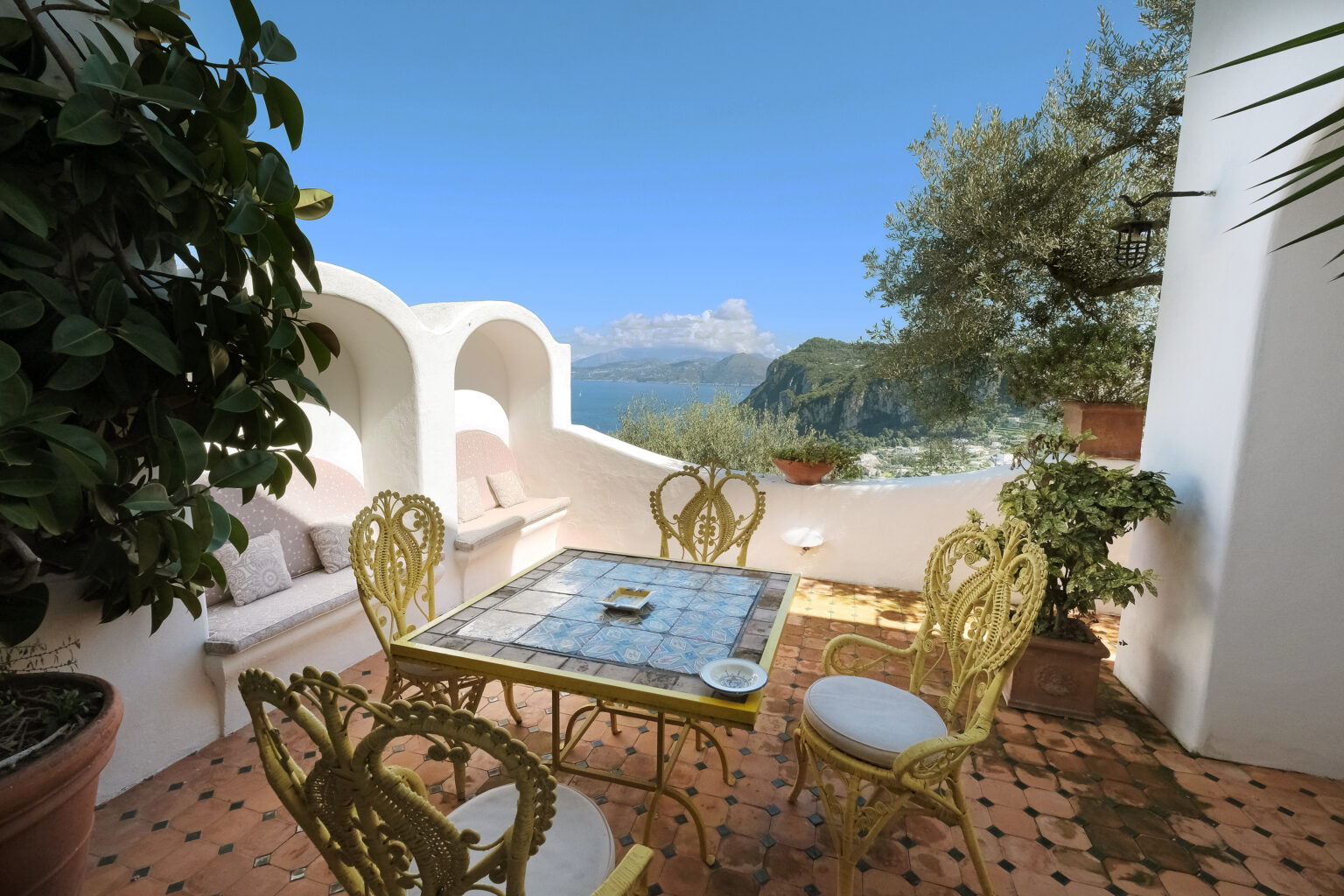 Cozy outdoor terrace with a stunning view, featuring a decorative gold table and chairs, surrounded by lush greenery and white walls, ideal for relaxation and dining. | Only on SopranoVillas