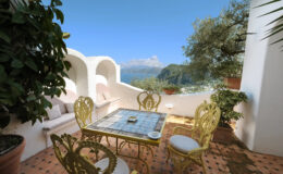 Cozy outdoor terrace with a stunning view, featuring a decorative gold table and chairs, surrounded by lush greenery and white walls, ideal for relaxation and dining.