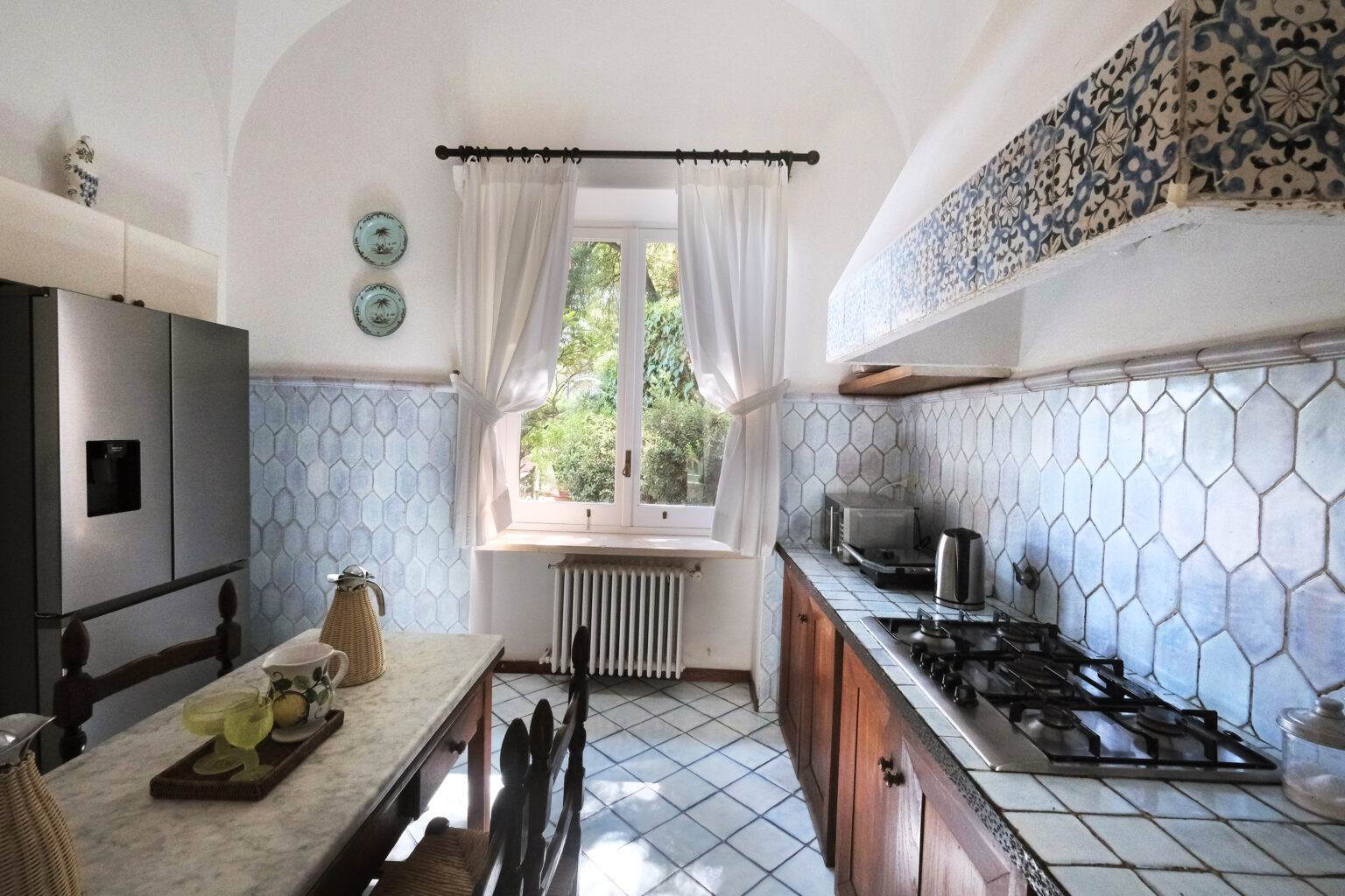 Bright and airy kitchen with blue hexagonal tiles, wooden cabinets, and a dining table. Natural light floods in through a window adorned with white curtains, creating a welcoming atmosphere. | Only on SopranoVillas