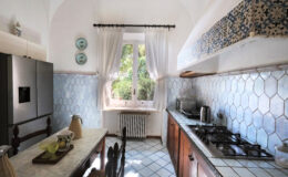 Bright and airy kitchen with blue hexagonal tiles, wooden cabinets, and a dining table. Natural light floods in through a window adorned with white curtains, creating a welcoming atmosphere.