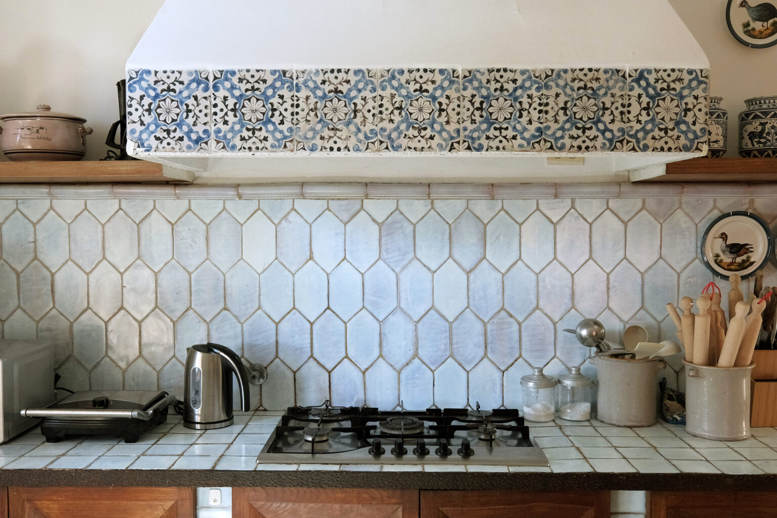 Rustic kitchen interior featuring a vintage gas stove, stainless steel kettle, and various cooking utensils on a tiled countertop. The backsplash showcases hexagonal light blue tiles and decorative patterned tiles above the stove, enhancing the warm, inviting atmosphere of the space. | Only on SopranoVillas