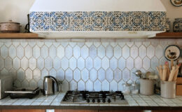 Rustic kitchen interior featuring a vintage gas stove, stainless steel kettle, and various cooking utensils on a tiled countertop. The backsplash showcases hexagonal light blue tiles and decorative patterned tiles above the stove, enhancing the warm, inviting atmosphere of the space.