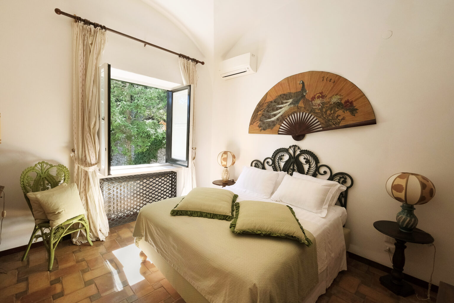 Cozy bedroom featuring a wrought iron bed with green accent pillows, a vintage fan art piece on the wall, and a charming green chair by a window that opens to a lush outdoor view. The room is illuminated by natural light, creating a warm and inviting atmosphere. | Only on SopranoVillas