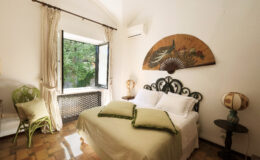 Cozy bedroom featuring a wrought iron bed with green accent pillows, a vintage fan art piece on the wall, and a charming green chair by a window that opens to a lush outdoor view. The room is illuminated by natural light, creating a warm and inviting atmosphere.