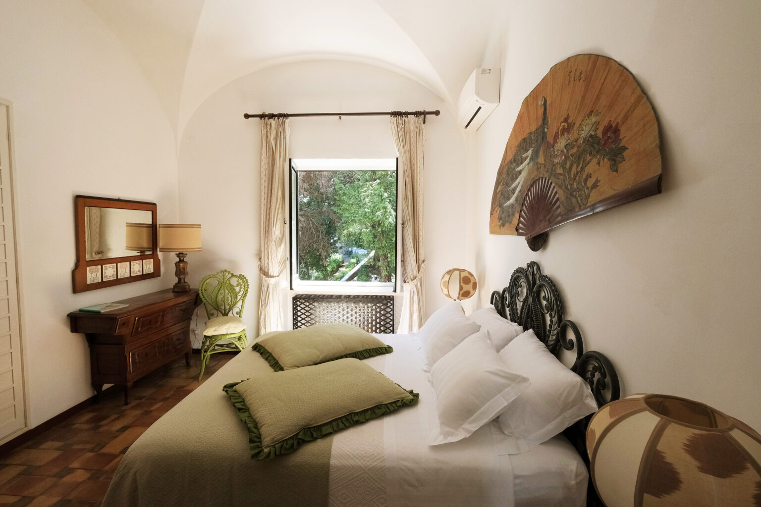 Cozy bedroom featuring a king-sized bed with green decorative pillows, vintage furniture, and a large window that offers a view of lush greenery. The room is adorned with a decorative wall fan and stylish lamps, creating a warm and inviting atmosphere. | Only on SopranoVillas