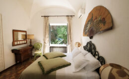 Cozy bedroom featuring a king-sized bed with green decorative pillows, vintage furniture, and a large window that offers a view of lush greenery. The room is adorned with a decorative wall fan and stylish lamps, creating a warm and inviting atmosphere.