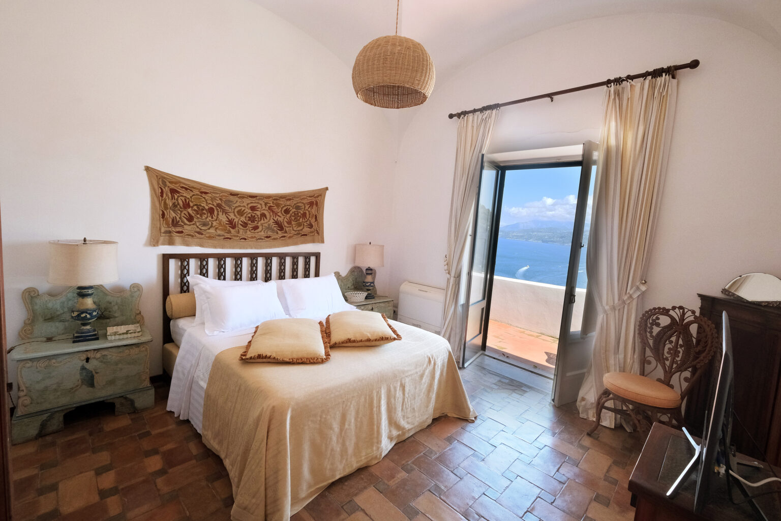 Cozy bedroom featuring a queen-sized bed with decorative pillows, a rustic wooden headboard, and an inviting view through large glass doors. The room is adorned with light-colored curtains, a woven wall hanging, and a vintage bedside table, creating a warm and tranquil atmosphere. | Only on SopranoVillas