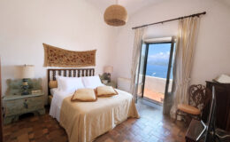 Cozy bedroom featuring a queen-sized bed with decorative pillows, a rustic wooden headboard, and an inviting view through large glass doors. The room is adorned with light-colored curtains, a woven wall hanging, and a vintage bedside table, creating a warm and tranquil atmosphere.