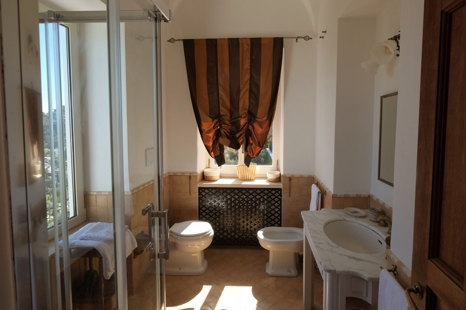 Spacious and elegantly designed bathroom featuring a glass shower, modern toilet fixtures, and decorative curtains. Natural light enhances the warm color palette and stylish decor. | Only on SopranoVillas