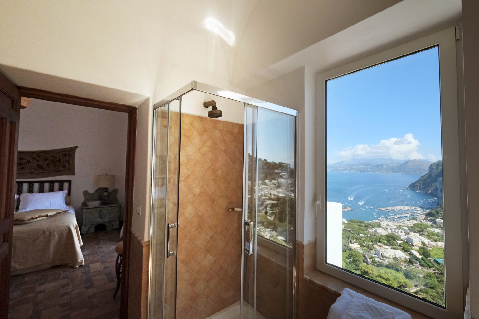 Modern bathroom with a glass shower enclosure and a stunning view of the coastline and mountains of Capri, Italy, visible through a large window. A cozy bedroom is seen in the background, enhancing the inviting atmosphere of the space. | Only on SopranoVillas