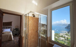 Modern bathroom with a glass shower enclosure and a stunning view of the coastline and mountains of Capri, Italy, visible through a large window. A cozy bedroom is seen in the background, enhancing the inviting atmosphere of the space.