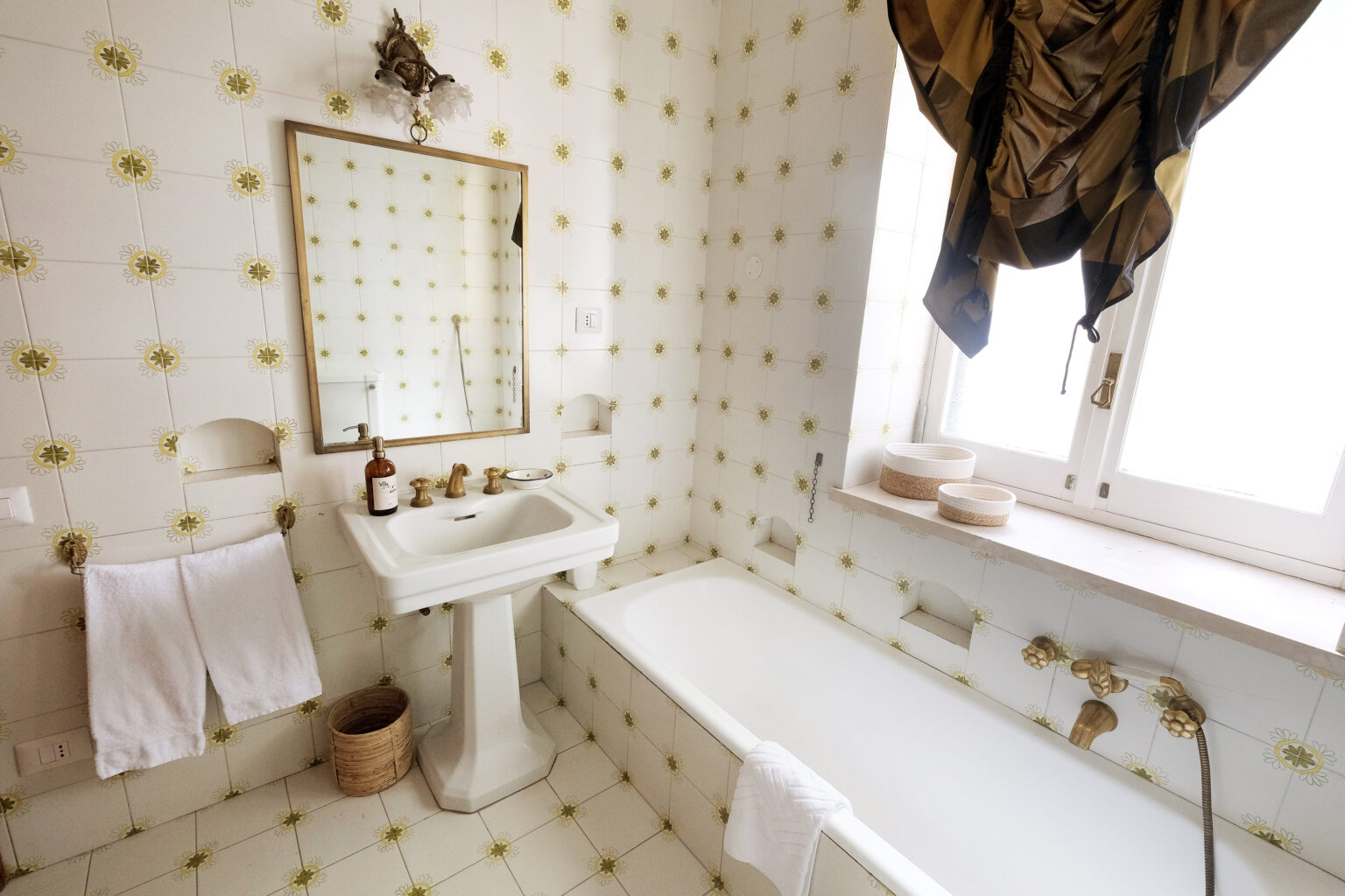 Bright and airy bathroom featuring a white bathtub, pedestal sink, and vintage-style mirror. The walls are adorned with yellow floral tiles, complemented by elegant drapes at the window and neatly hung towels. | Only on SopranoVillas
