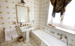Bright and airy bathroom featuring a white bathtub, pedestal sink, and vintage-style mirror. The walls are adorned with yellow floral tiles, complemented by elegant drapes at the window and neatly hung towels.