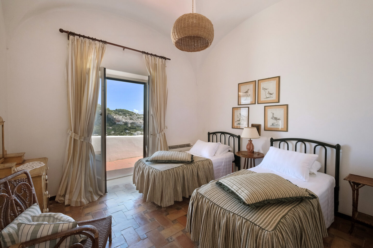 Cozy bedroom featuring two twin beds with green and white bedding, wooden furniture, and a large window with scenic views. Natural light illuminates the space, complemented by decorative wall art and a woven light fixture. Ideal for a relaxing getaway. | Only on SopranoVillas