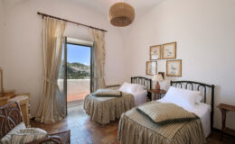 Cozy bedroom featuring two twin beds with green and white bedding, wooden furniture, and a large window with scenic views. Natural light illuminates the space, complemented by decorative wall art and a woven light fixture. Ideal for a relaxing getaway.