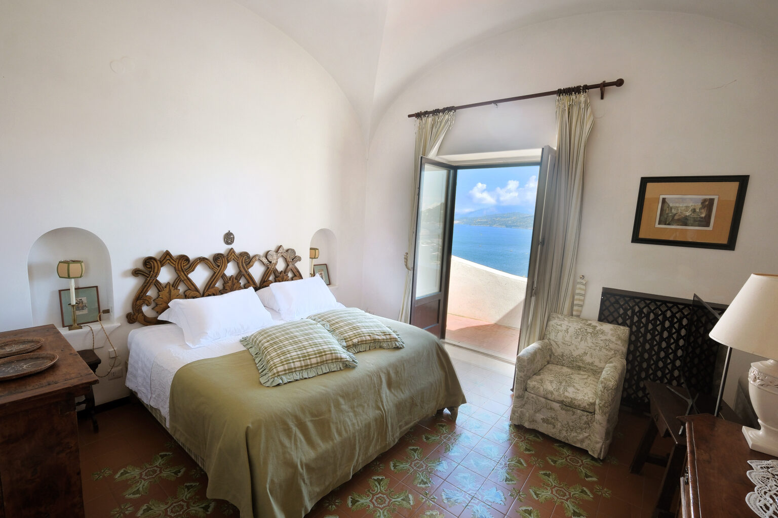 Cozy bedroom with a large bed, decorative headboard, and a chair, featuring a stunning ocean view through an open door. The room is bright and airy, with light-colored walls and elegant furnishings, ideal for relaxation. | Only on SopranoVillas