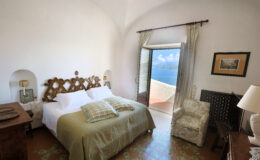 Cozy bedroom with a large bed, decorative headboard, and a chair, featuring a stunning ocean view through an open door. The room is bright and airy, with light-colored walls and elegant furnishings, ideal for relaxation.